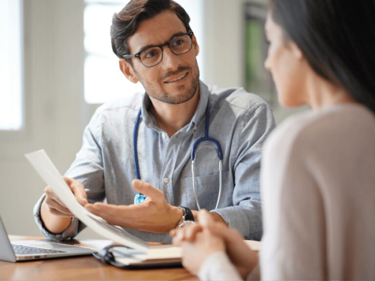 Man wearing stethoscope gesturing to document and speaking with woman