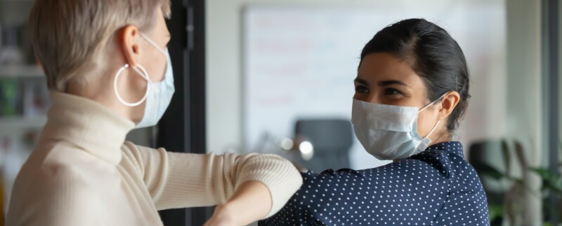 Two office workers wearing facial masks bump elbows in greeting.