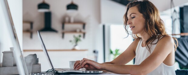 woman in white tank top seated at kitchen table completing coursework on laptop
