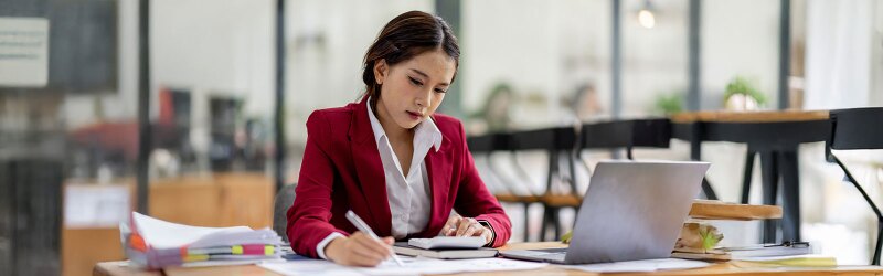 Business admin working on laptop, using financial calculator at modern office, analyzing business document with financial graph.