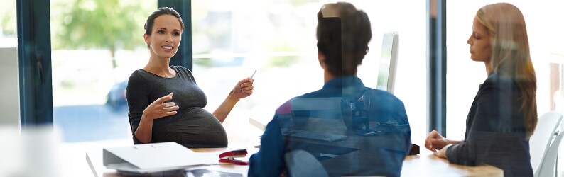A pregnant woman is sitting at a table in a discussion with two coworkers.