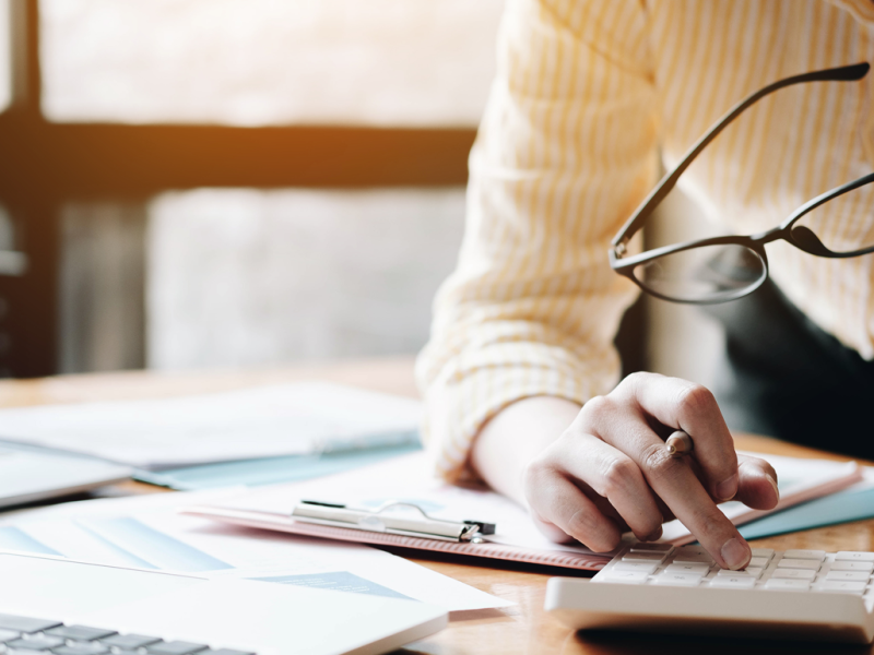 A woman sitting at a desk, holding a pair of glasses and typing on a calculator.