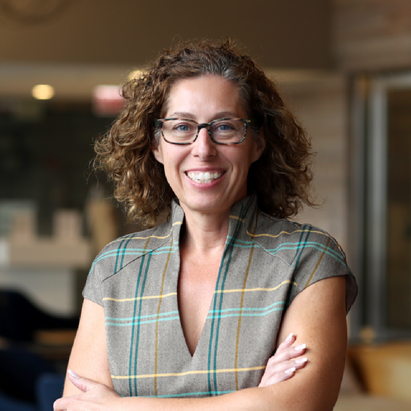 Headshot of Teelina House with curly hair and glasses, arms crossed, wearing a plaid top in a softly lit indoor setting.