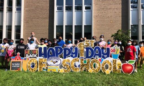 Haley Padgett and her fifth grade students stand outside of their school in front of a large Happy First Day of School sign