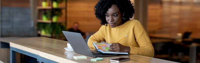 A woman in a cafe setting, focused on her laptop and notebook, searching for job opportunities