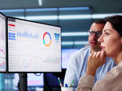 Professionals are gathered around two monitors displaying various business analytics charts and graphs, with one woman thoughtfully observing the data.