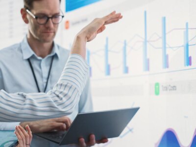 Caucasian male and female data scientists with laptop and tablet standing next to big digital screen with graphs and charts in monitoring office.
