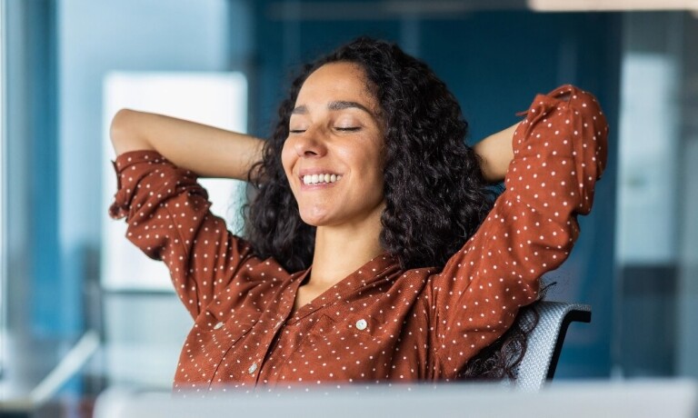 Woman sits at office table with hands behind her head, eyes closed
