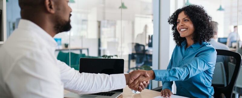 Man and woman shake hands across a desk after an interview.