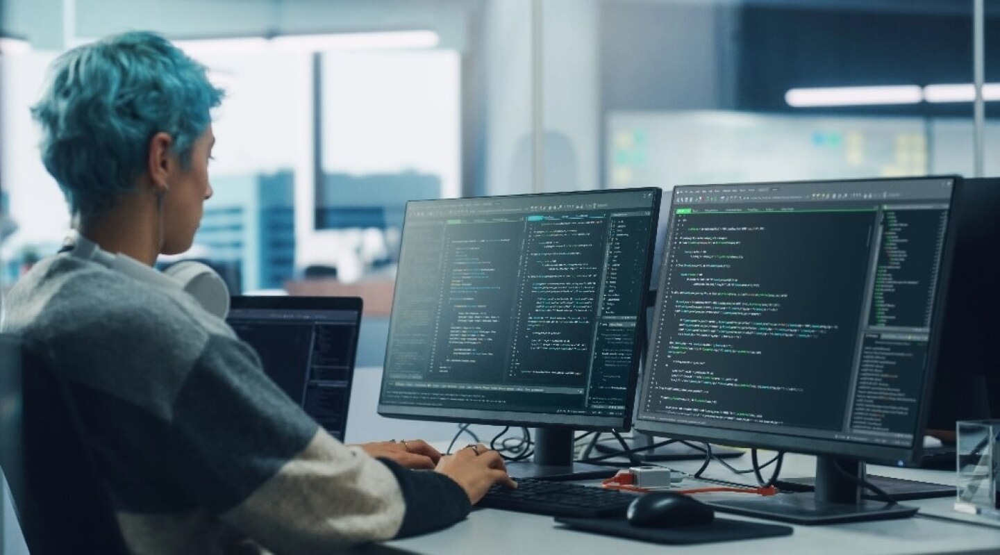 Data Scientist in casual clothing with blue hair writing code on multiple displays