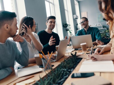 Group of young professionals in smart casual wear discussing something and smiling while seated around a table, with laptop computers