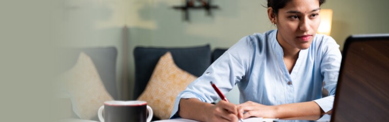 Woman wearing light blue tunic holds pen while looking at a laptop screen in living room