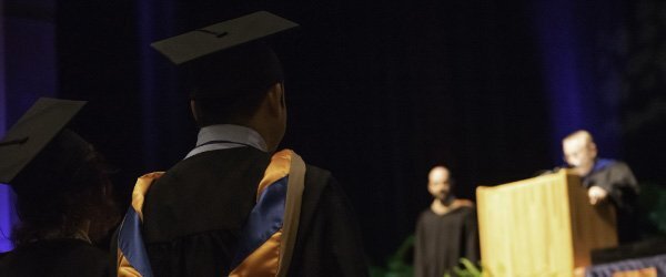 two-MBA-graduates-in-caps-and-gowns-with-backs-to-viewer-and-podium-and-graduation-speaker-in-foreground