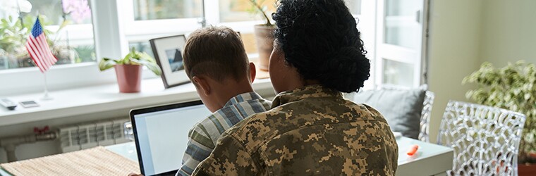 Military mother sits at dining room table with child on her lap while working on a laptop