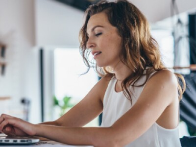 woman in white tank top seated at kitchen table completing coursework on laptop