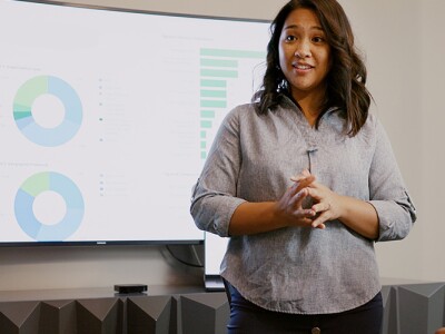 Woman stands in front of a group of her peers, presenting information depicted on a screen behind her.