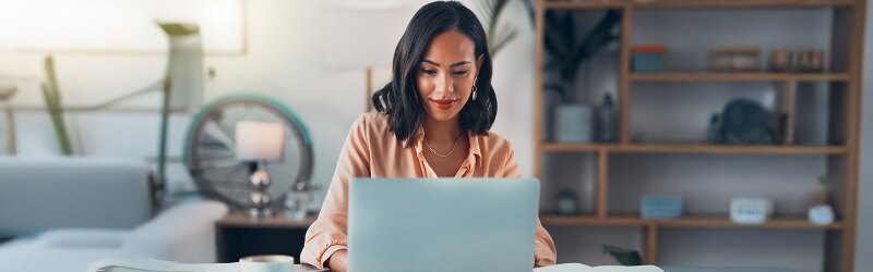 A smiling woman with dark hair sits at a desk, looking down at a laptop. A blurred room is in the background.