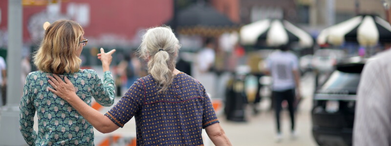Two women walk together through a New York City neighborhood, talking as one gestures ahead.