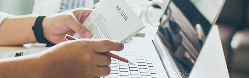 Hands holding a resume and a red pen while resting on a desk with a laptop