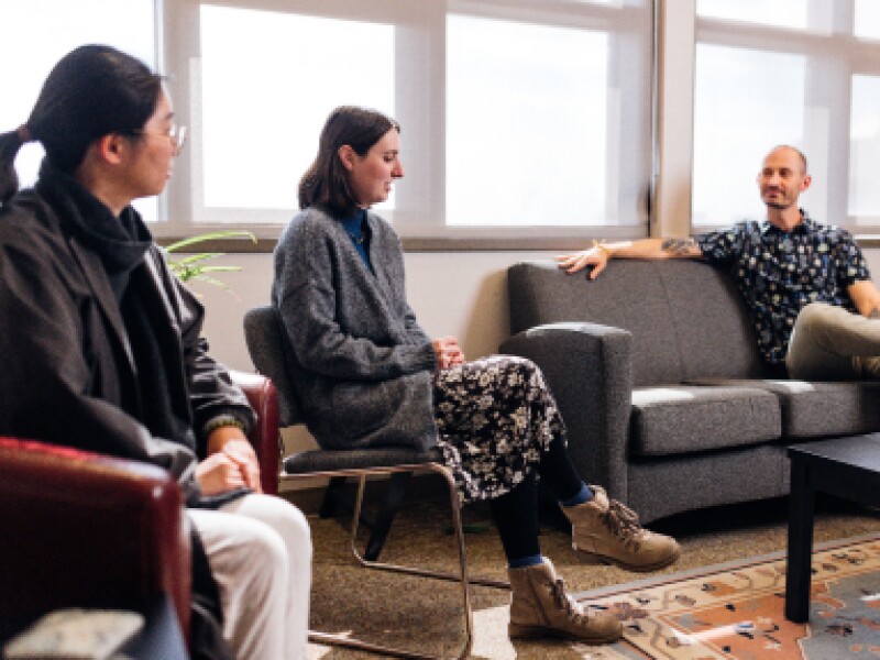 Two female KU alumni are talking with a male KU student sitting on a couch.