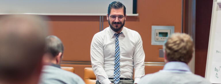 Professional in white button up shirt and tie sits in front of an audience of people