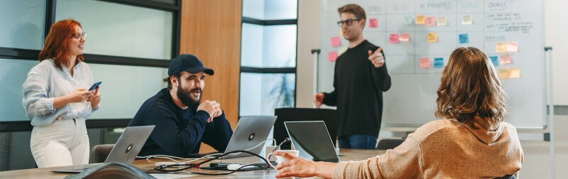 A person is leading a discussion at a whiteboard with sticky notes, while others participate in conversation