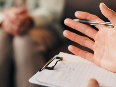 A therapist takes notes in the foreground while a patient sits in the background
