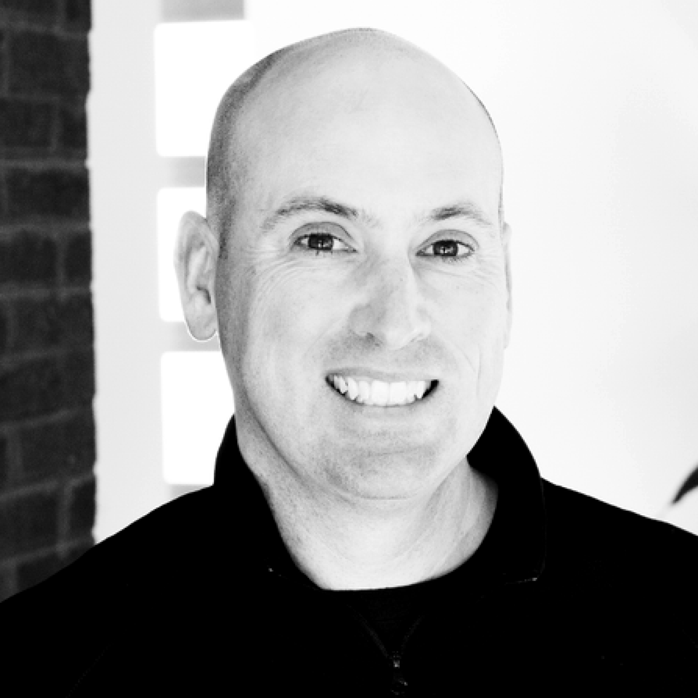 Black-and-white headshot of a smiling bald man in a dark zip-up jacket, standing indoors with soft light and a blurred background.