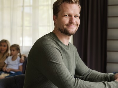 Smiling man working on laptop at a desk. His wife and daughter are watching tv on sofa in background