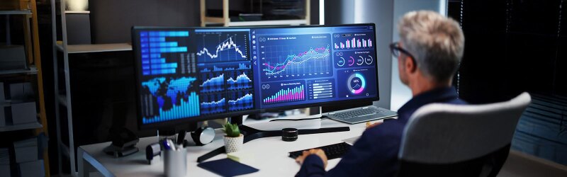 A man analyzing data on multiple monitors displaying charts, graphs, and global business metrics in a modern office setting.