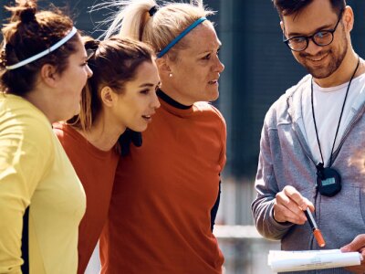 Female players and their coach going through game strategy before soccer match on playing field.