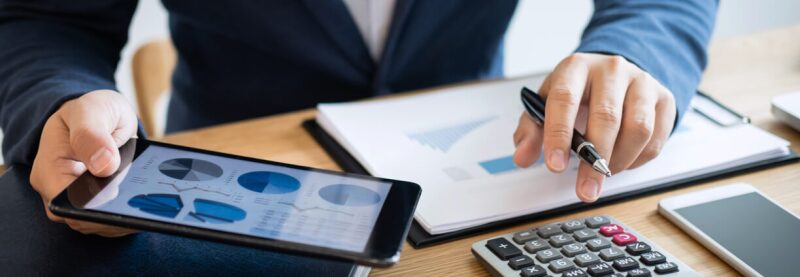 Hands of accountant in suit using tablet with charts, clipboard and calculator at desk