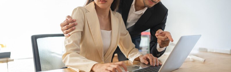 woman seated at desk looks uncomfortable as man wraps arms around her pointing at her laptop screen