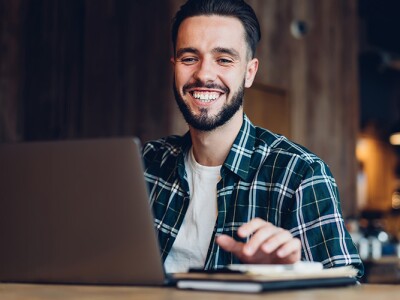 Smiling man in casual attire working on a laptop in a warmly lit cafe.