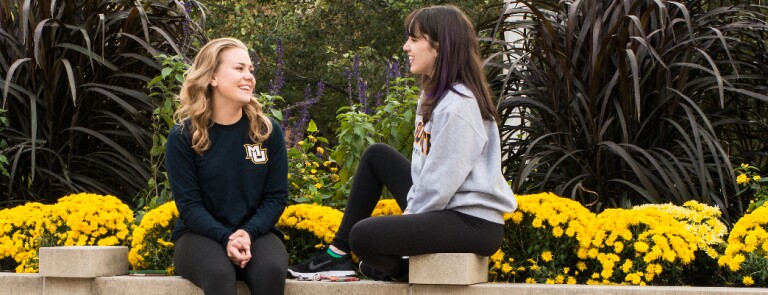 Two female Marquette business students talking on a bench near a garden
