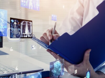 Person in lab coat with an open blue folder and pen in hand standing in front of a laptop