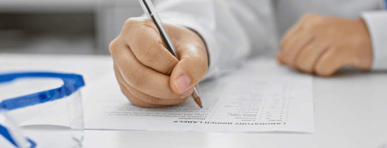 Hands of clinical research professional filling out form near microscope and lab goggles