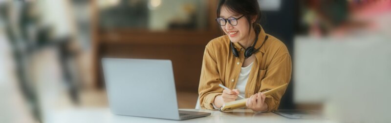 Asian girl student reviews her college application status on her laptop while taking notes.