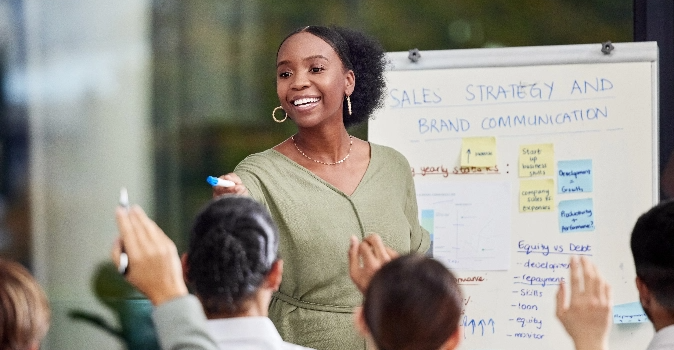 Woman at white board in front of room of colleagues raising hands