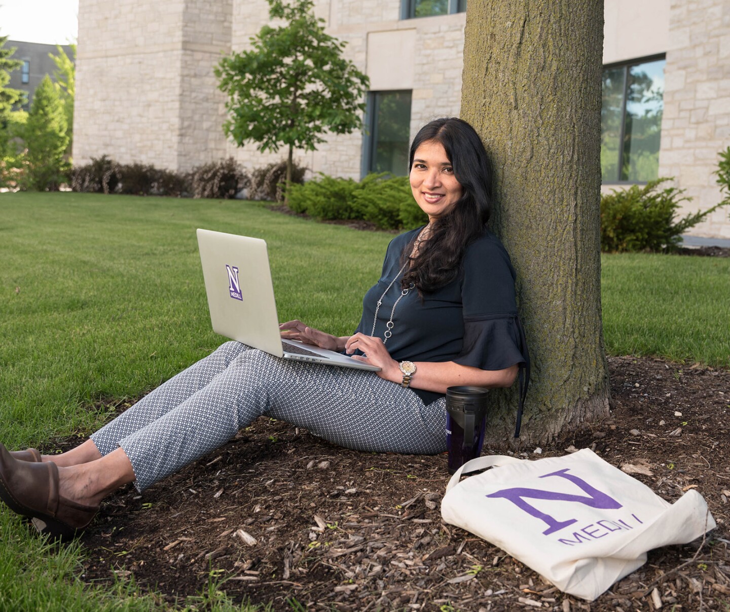 Woman sitting under a tree with a laptop and a Medill tote bag, smiling at the camera.