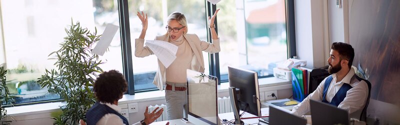 A woman stands at the center of an office surrounded by two seated colleagues. The woman seems angry and is tossing papers into the air.