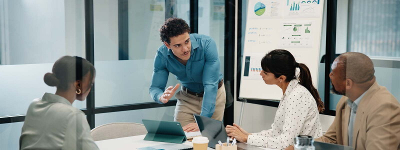 A business team meeting around a table in a modern office near a whiteboard and laptops.