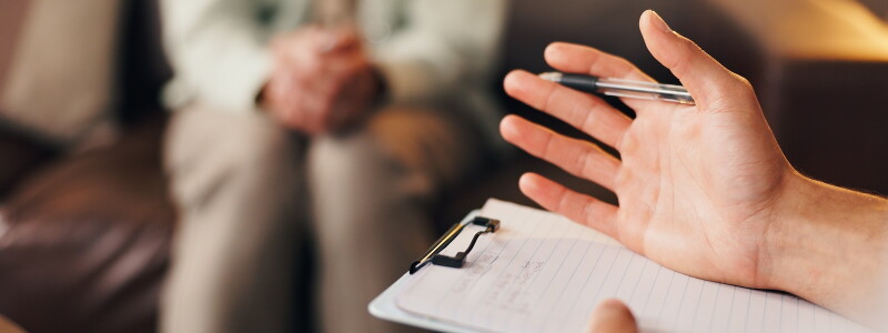 A therapist takes notes in the foreground while a patient sits in the background